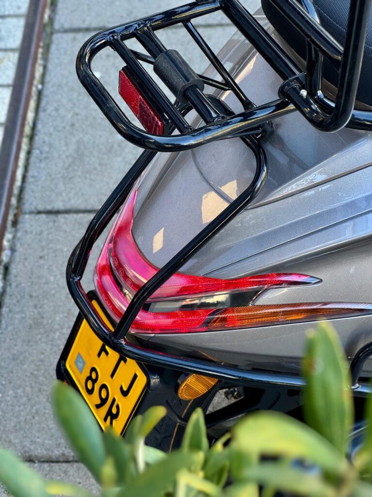 Achterkant van een grijze scooter met bagagedrager, achterlicht en geel kenteken.