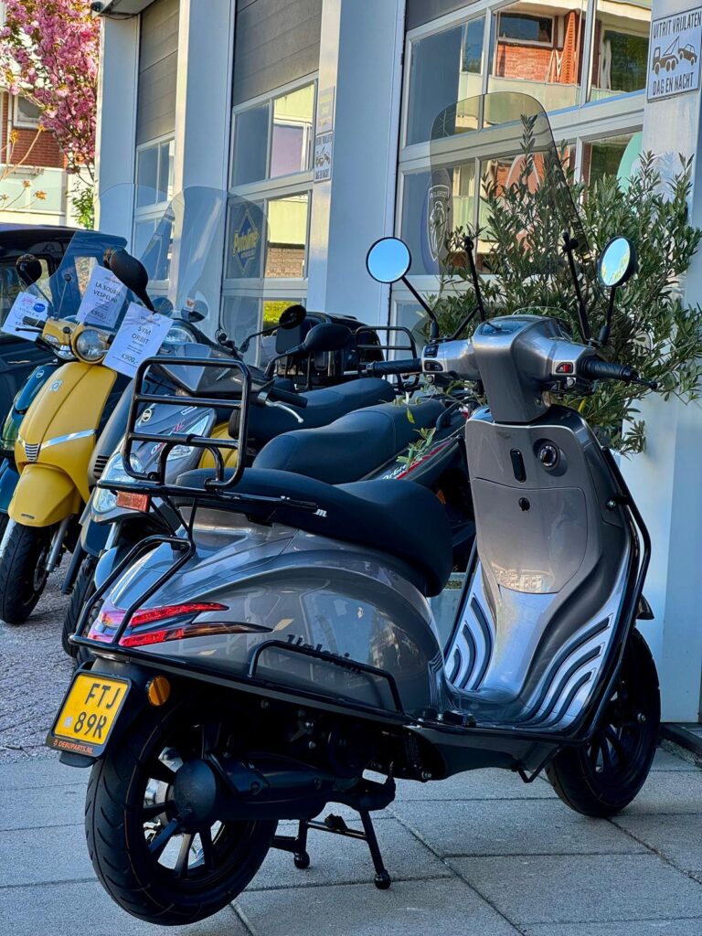 Grijze scooter met bagagerek geparkeerd voor een scootershop, met meerdere scooters op de achtergrond.