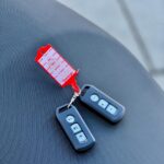 Twee zwarte afstandsbedieningen met rode labelsleutelhanger op een grijs scooterzadel.