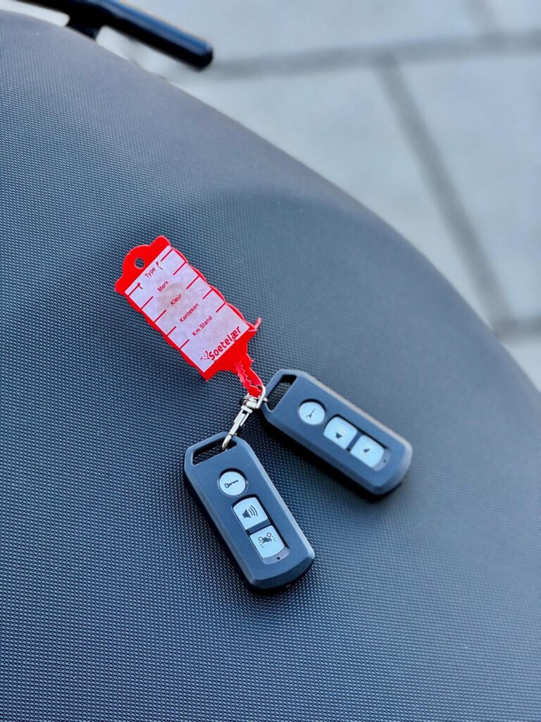 Twee zwarte afstandsbedieningen met rode labelsleutelhanger op een grijs scooterzadel.