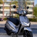 Grijze scooter met hoog windscherm en bagagedrager, geparkeerd op stoep voor appartementen.
