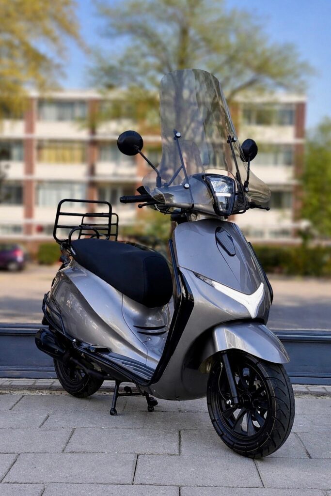 Grijze scooter met hoog windscherm en bagagedrager, geparkeerd op stoep voor appartementen.