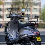 Grijze scooter met hoog windscherm en bagagedrager, geparkeerd op stoep voor flats.