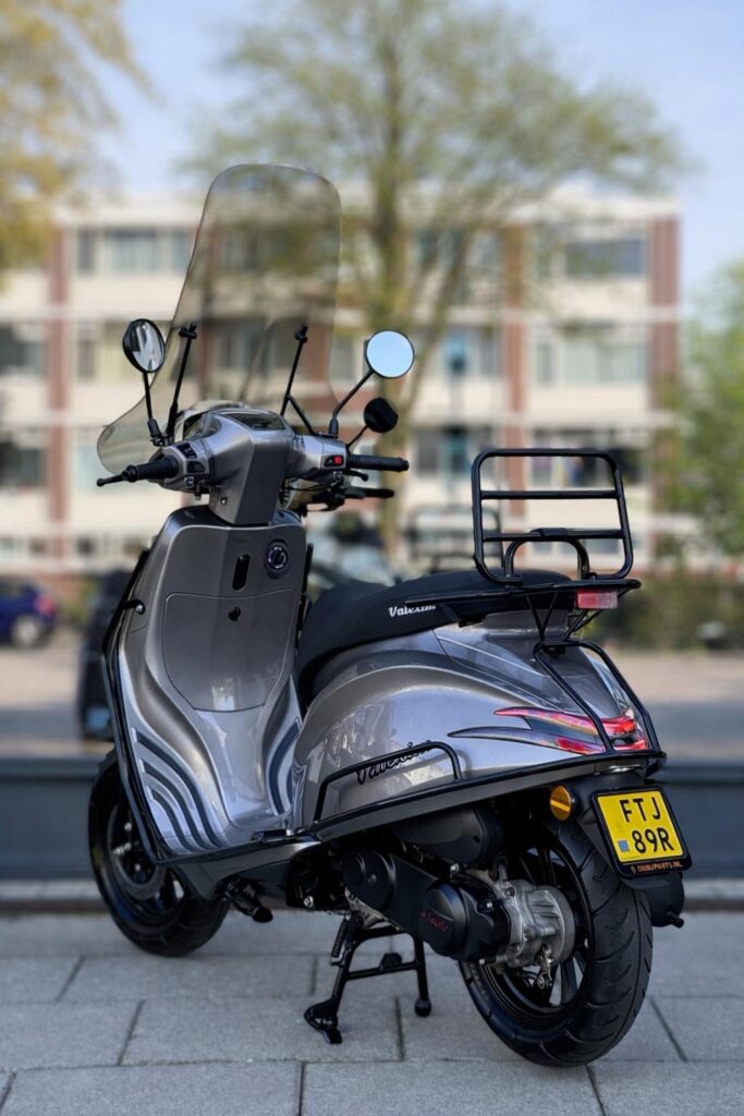 Grijze scooter met hoog windscherm en bagagedrager, geparkeerd op stoep voor flats.