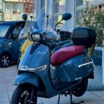 Blauwgrijze retro scooter met bordeauxrood zadel, topkoffer en hoog windscherm, geparkeerd bij showroom.