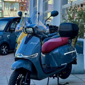 Blauwgrijze retro scooter met bordeauxrood zadel, topkoffer en hoog windscherm, geparkeerd bij showroom.