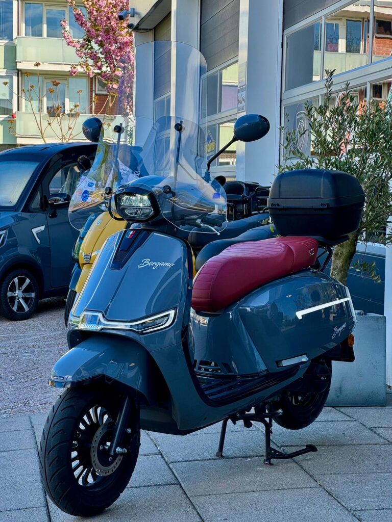 Blauwgrijze retro scooter met bordeauxrood zadel, topkoffer en hoog windscherm, geparkeerd bij showroom.