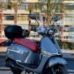 Grijze retro-scooter met rood zadel, windscherm en topkoffer, geparkeerd voor etalage.