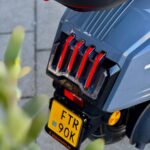 Achterkant van een grijze scooter met rode LED-achterlichten en een gele NL-kentekenplaat.