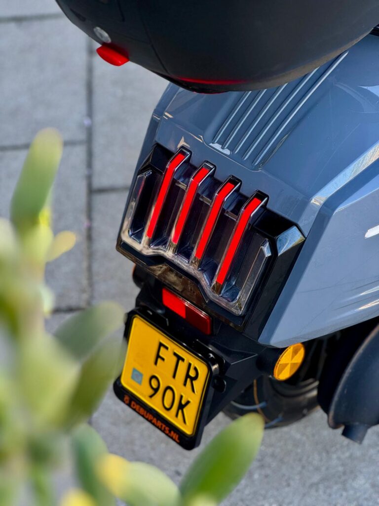 Achterkant van een grijze scooter met rode LED-achterlichten en een gele NL-kentekenplaat.