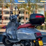 Grijze scooter met rood zadel en topkoffer, geparkeerd op de stoep voor een raam met flat in reflectie.