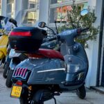 Grijze scooter met rood zadel en topkoffer, geparkeerd voor een scooterzaak naast een gele scooter.