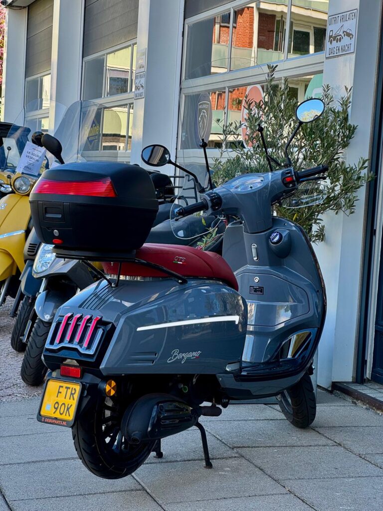 Grijze scooter met rood zadel en topkoffer, geparkeerd voor een scooterzaak naast een gele scooter.