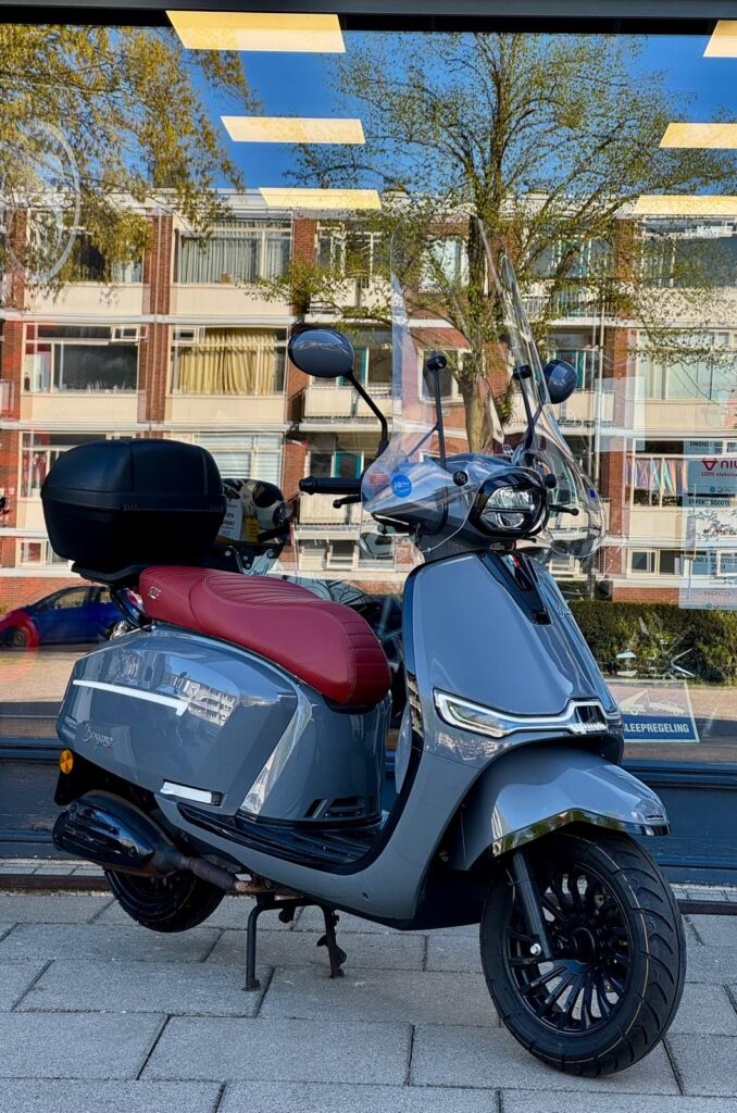 Grijze retro-scooter met rood zadel, windscherm en topkoffer, geparkeerd voor etalage.