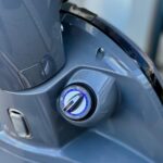 Close-up van het contactslot van een grijze scooter met blauwe ring.