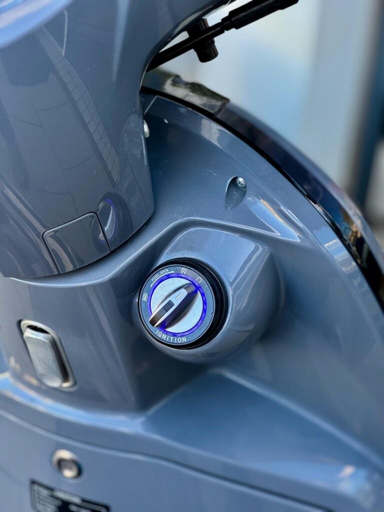 Close-up van het contactslot van een grijze scooter met blauwe ring.