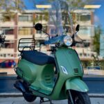 Matgroene retro-scooter met windscherm op standaard, voor een raam met weerspiegelde flats.