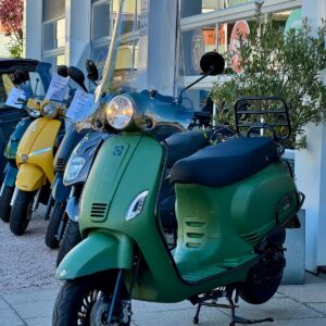 Matgroene scooter met hoog windscherm op de standaard, geparkeerd voor een showroom.