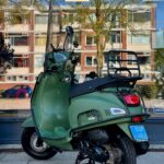 Groene scooter met hoog windscherm op middenstandaard, geparkeerd voor etalageruit met flats en boom.