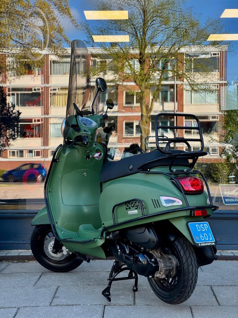 Groene scooter met hoog windscherm op middenstandaard, geparkeerd voor etalageruit met flats en boom.