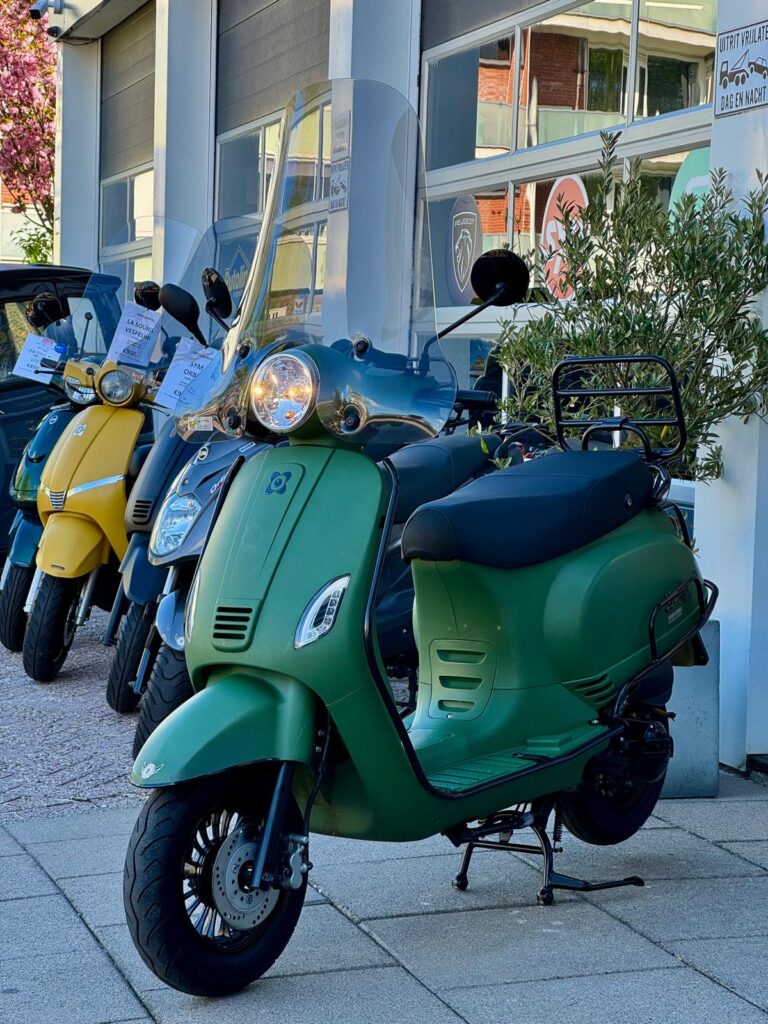 Matgroene scooter met hoog windscherm op de standaard, geparkeerd voor een showroom.