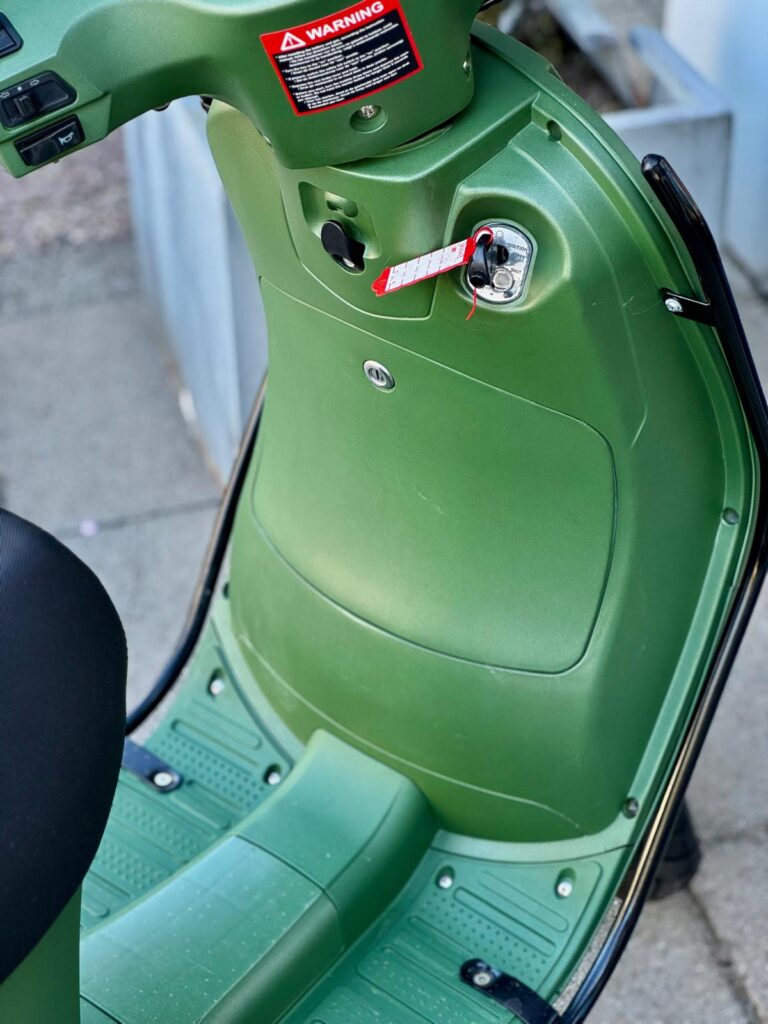 Detail van groene scooter: voetplaat, opbergvak en contactslot met label.