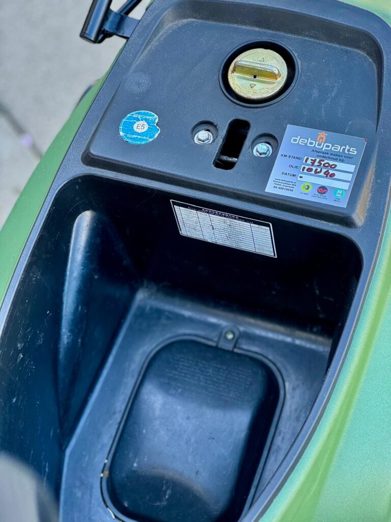 Close-up van scooteropbergruimte onder het zadel met brandstofdop en onderhoudssticker.