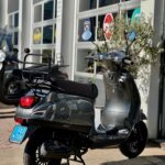 Donkergrijze AGM VX50 scooter met windscherm, geparkeerd voor een showroom met glazen roldeuren.