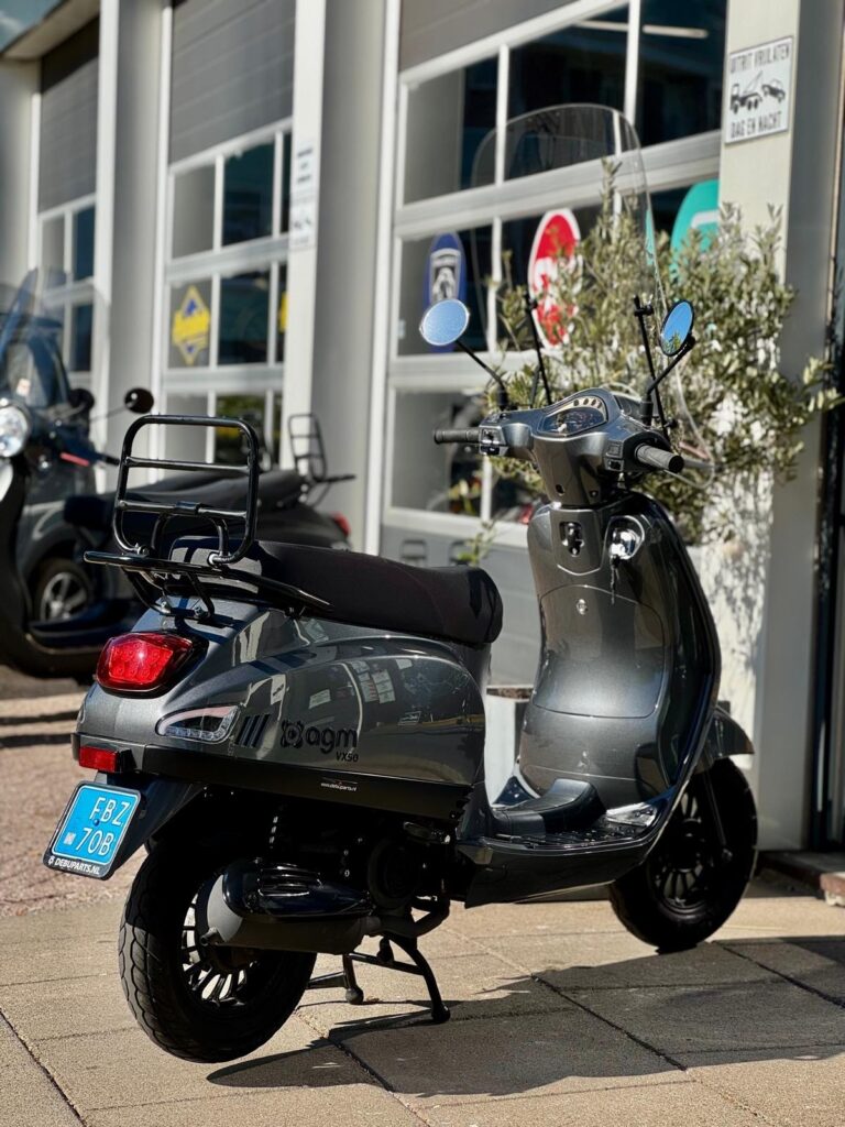 Donkergrijze AGM VX50 scooter met windscherm, geparkeerd voor een showroom met glazen roldeuren.