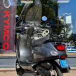 Donkergrijze scooter met windscherm en bagagerek, geparkeerd voor een winkel.