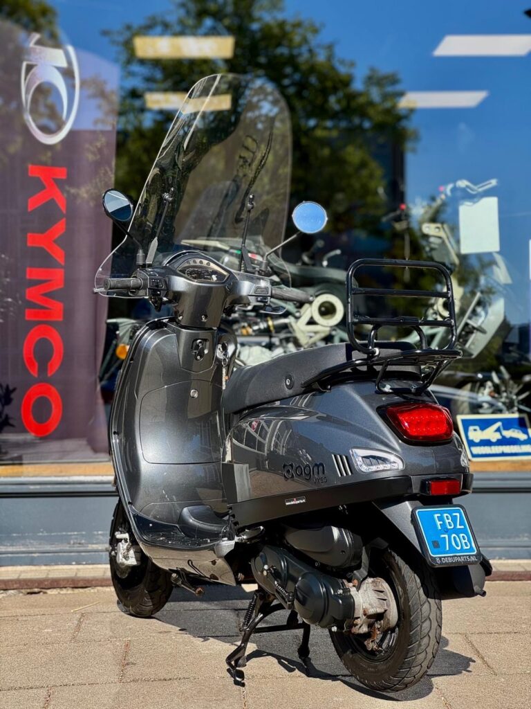 Donkergrijze scooter met windscherm en bagagerek, geparkeerd voor een winkel.