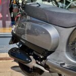 Close-up van de zijkant van een grijze AGM VX50 scooter met bagagedrager en uitlaat, geparkeerd bij een winkel.