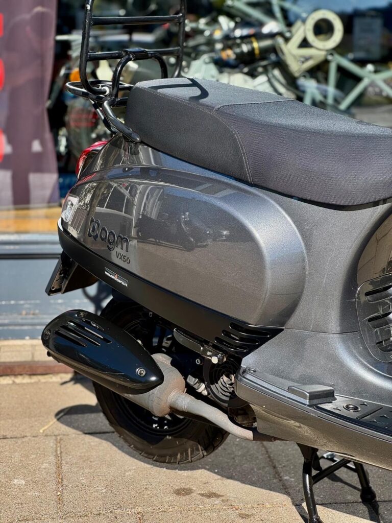 Close-up van de zijkant van een grijze AGM VX50 scooter met bagagedrager en uitlaat, geparkeerd bij een winkel.