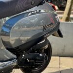 Close-up van de grijze AGM VX50 scooter met zadel, motorblok en achterwiel, geparkeerd op stoep.