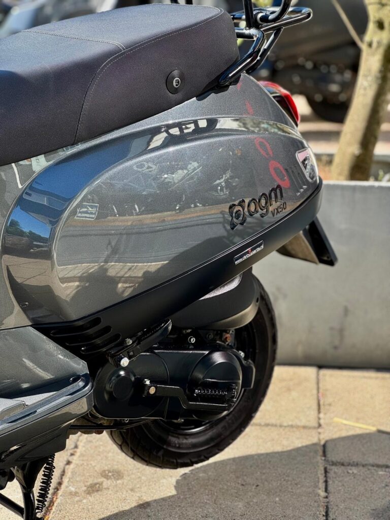 Close-up van de grijze AGM VX50 scooter met zadel, motorblok en achterwiel, geparkeerd op stoep.