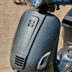 Close-up van de voorkant van een grijs metallic scooter met ronde koplamp en LED-dagrijverlichting in de zon.
