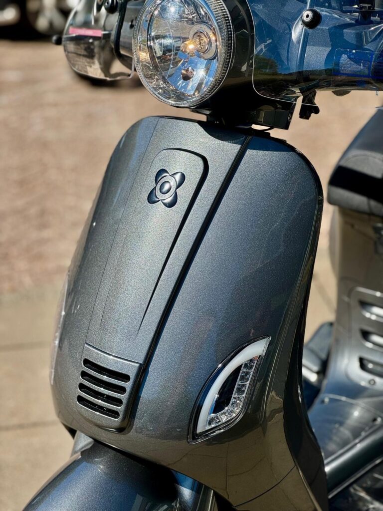 Close-up van de voorkant van een grijs metallic scooter met ronde koplamp en LED-dagrijverlichting in de zon.