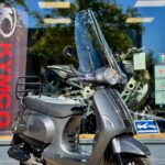 Donkergrijze scooter met hoog windscherm op de middenbok voor een showroom met KYMCO-banner.