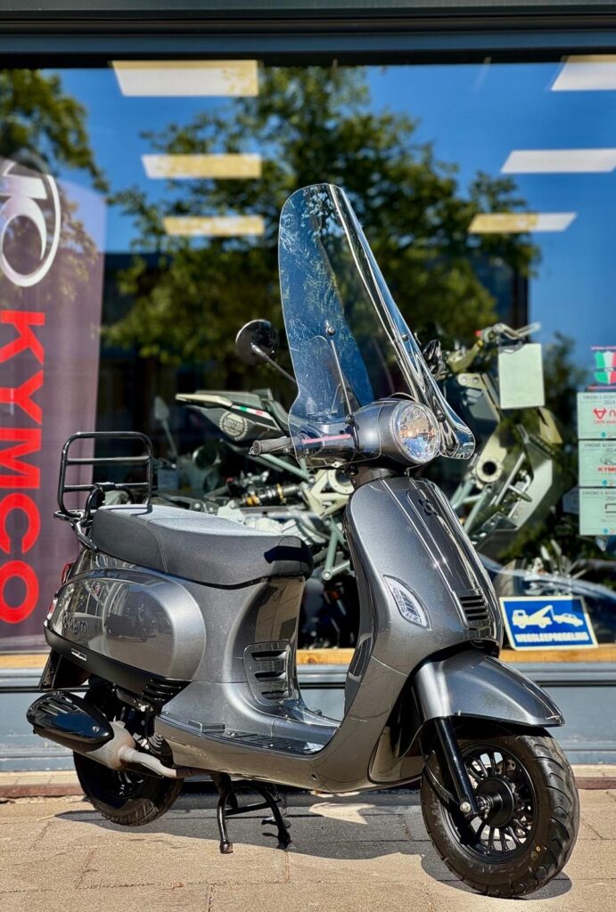 Donkergrijze scooter met hoog windscherm op de middenbok voor een showroom met KYMCO-banner.