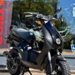 Zwarte Peugeot-scooter met grove offroadbanden voor een showroomraam in de zon.