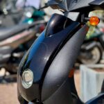 Close-up van de voorkant van een zwarte scooter met koplamp en oranje knipperlichten, buiten in de zon.