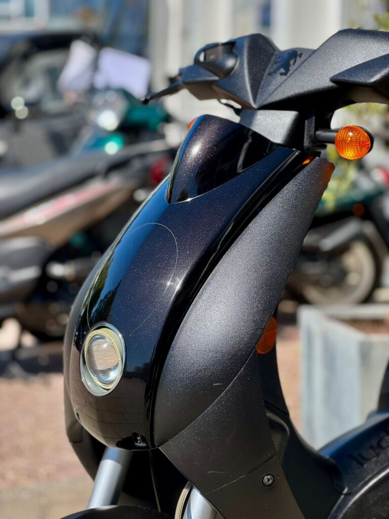 Close-up van de voorkant van een zwarte scooter met koplamp en oranje knipperlichten, buiten in de zon.