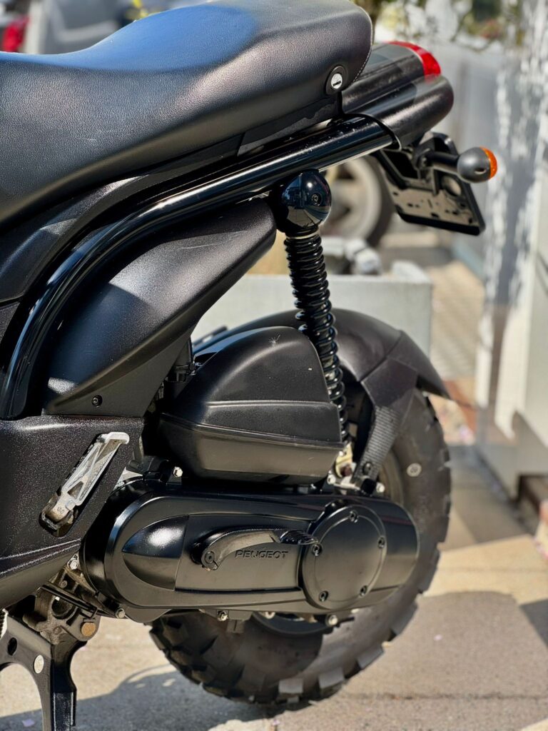 Close-up van de achterzijde van een zwarte Peugeot-scooter met vering, kentekenhouder en noppenband.