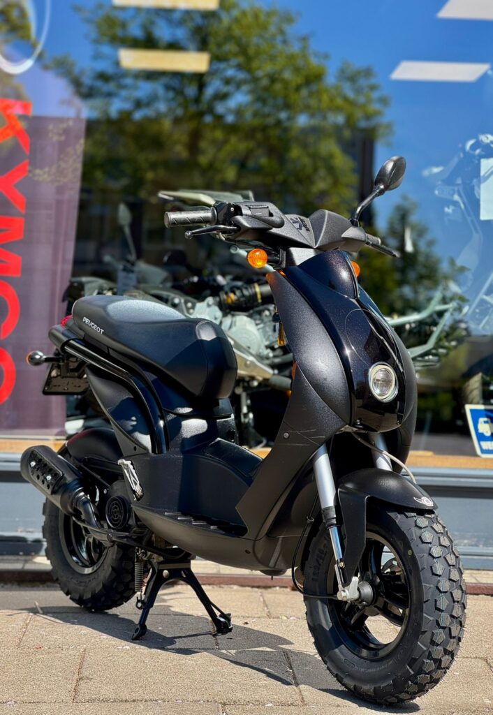 Zwarte Peugeot-scooter met grove offroadbanden voor een showroomraam in de zon.