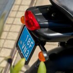 Achterkant van een zwarte scooter met rood achterlicht, oranje knipperlichten en blauwe kentekenplaat in de zon.