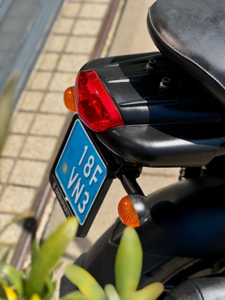 Achterkant van een zwarte scooter met rood achterlicht, oranje knipperlichten en blauwe kentekenplaat in de zon.