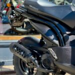 Close-up van de achterzijde van een zwarte Peugeot scooter met noppenband en uitlaat, geparkeerd bij een etalage.