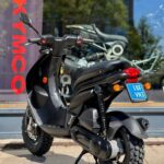 Zwarte Kymco-scooter met offroad-banden, op standaard geparkeerd voor een winkelruit met showroom.