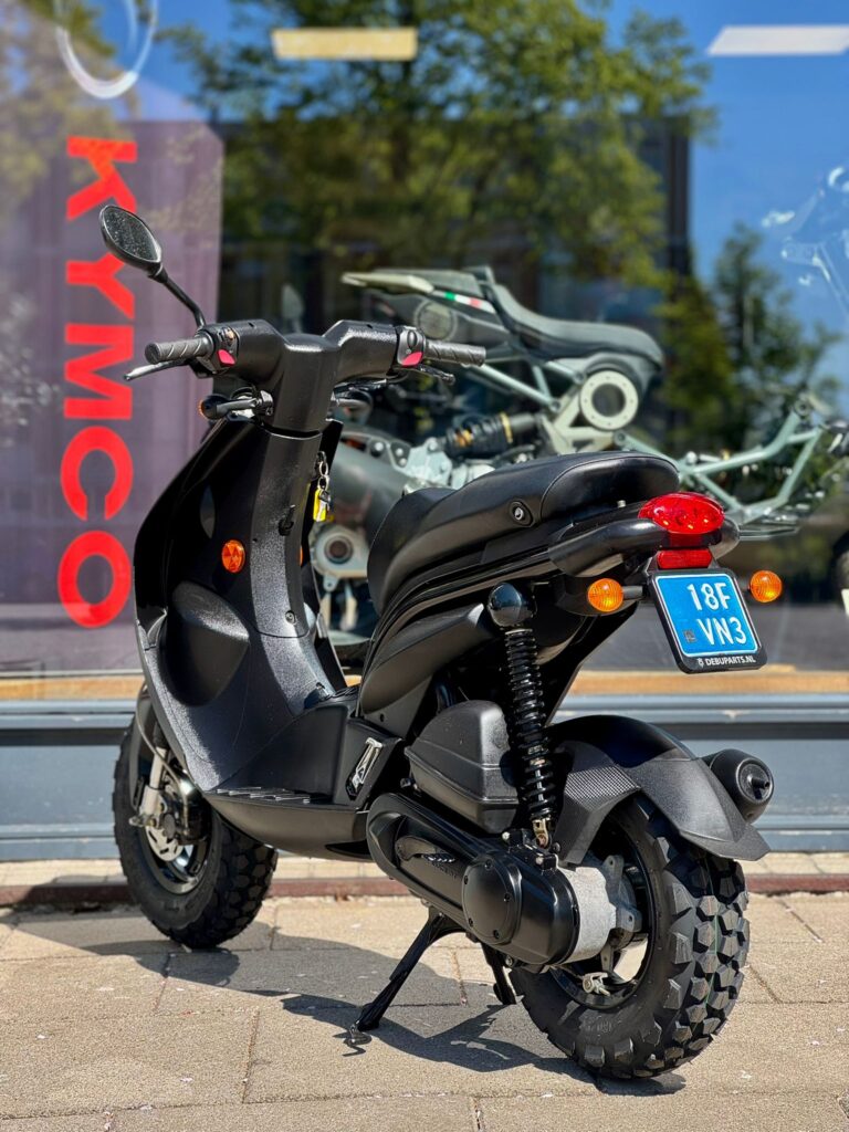 Zwarte Kymco-scooter met offroad-banden, op standaard geparkeerd voor een winkelruit met showroom.