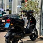 Zwarte Peugeot-scooter met grove terreinbanden geparkeerd voor een scooterwinkel in de zon.